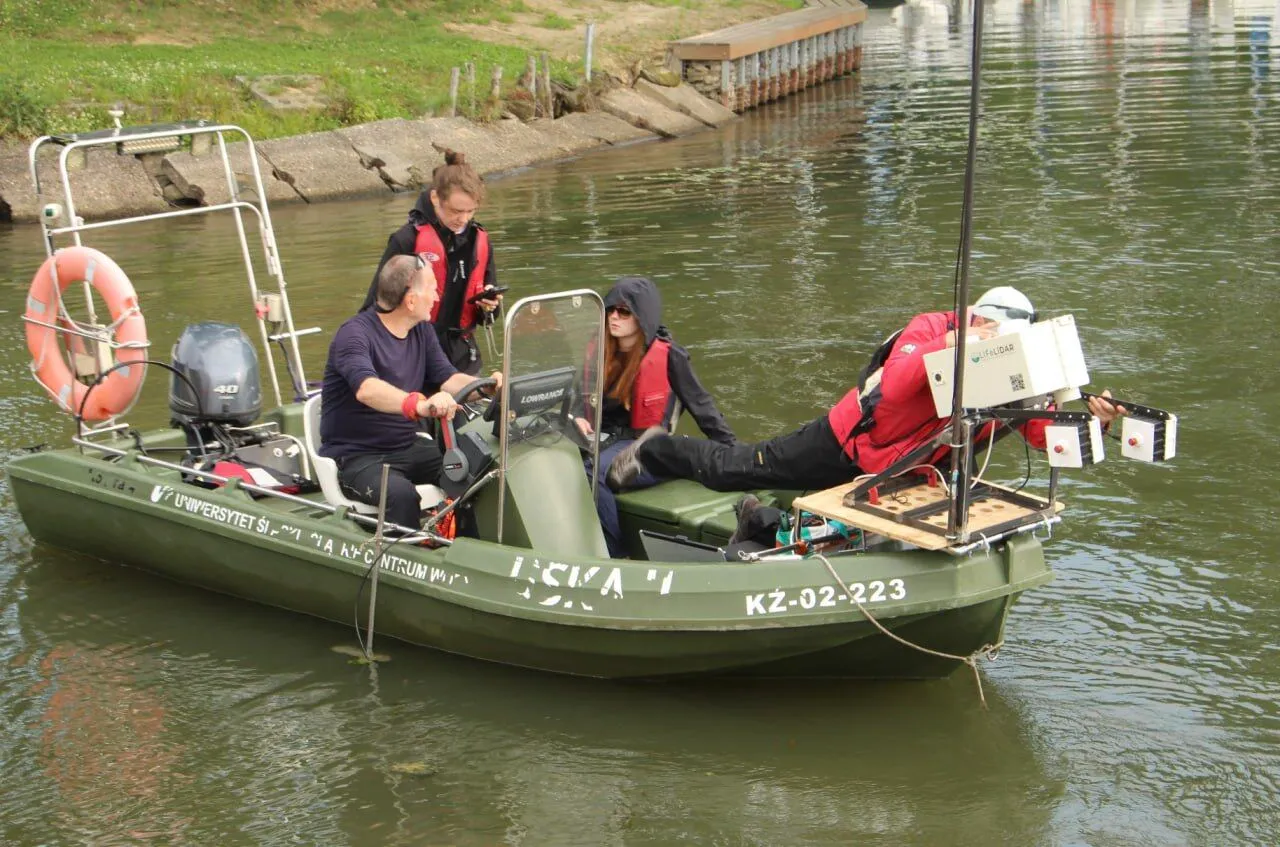 Four people aboard a green survey boat, the lidar rig mounted on a mast at the stern, a reed bank behind